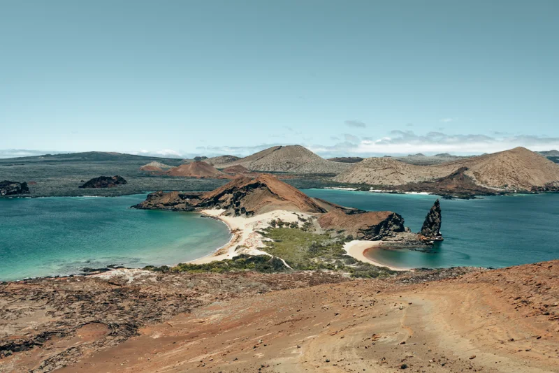 Looking out from a red volcanic hill over two turquoise bays and a tall pinnacle of rock.