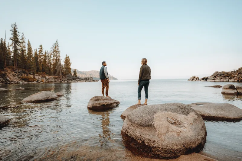 Two people standing on boulders at the edge of a clear lake, pine trees on the far shore.