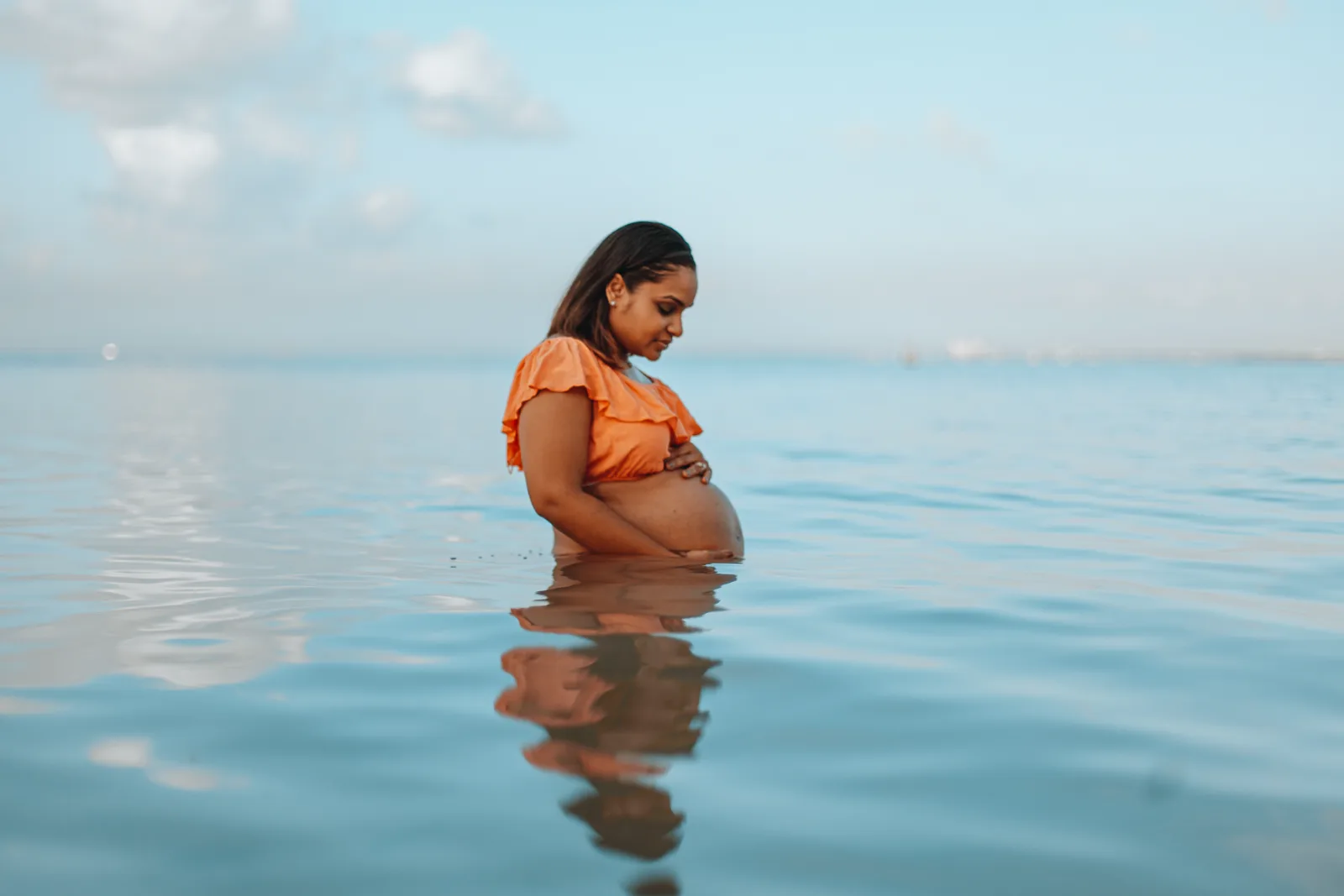 An expectant mother in an orange top stands in calm, shallow water, looking down at her belly.